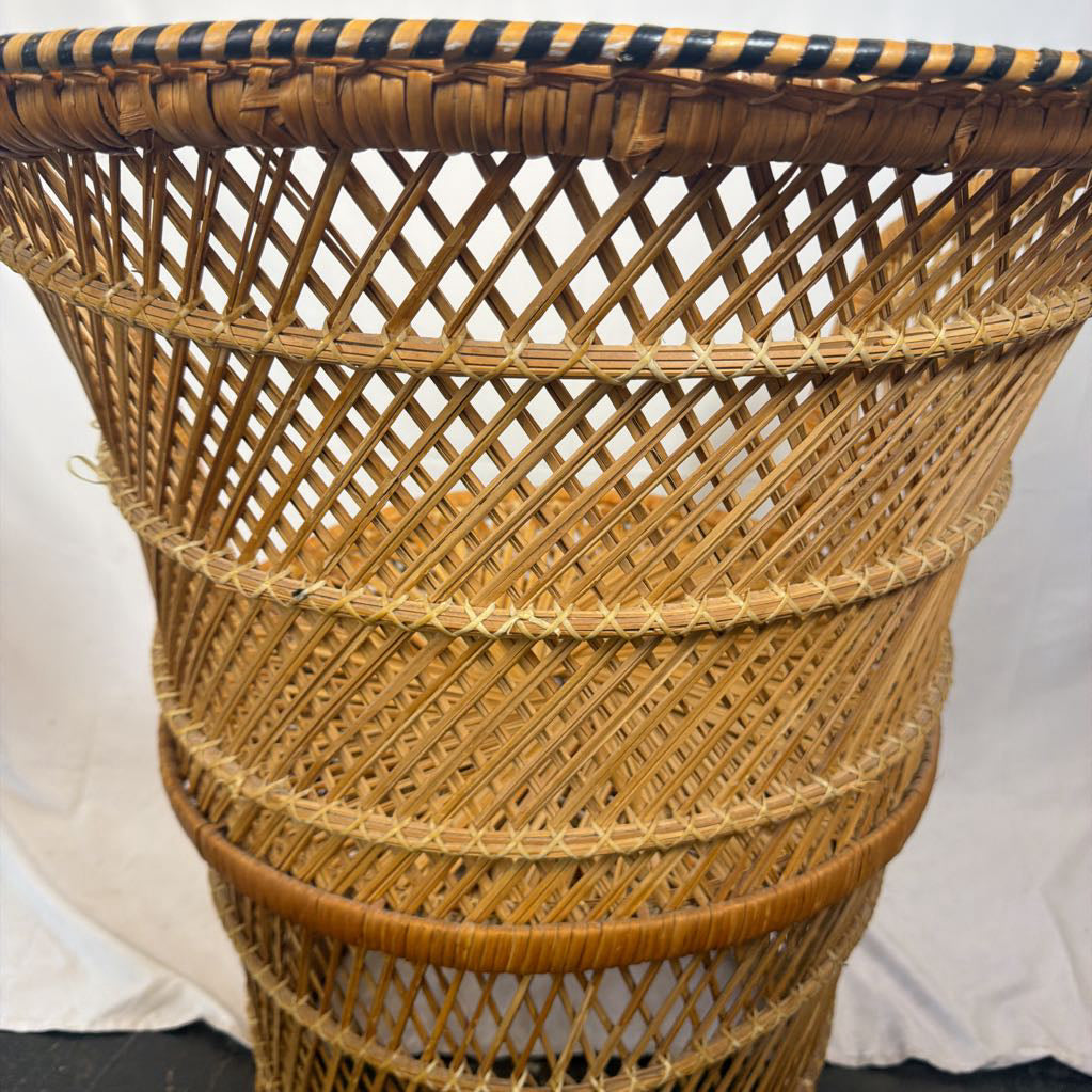 A natural woven rattan basket with a conical shape, featuring a diamond lattice pattern and a dark striped woven trim along the rim, set against a white fabric backdrop.
