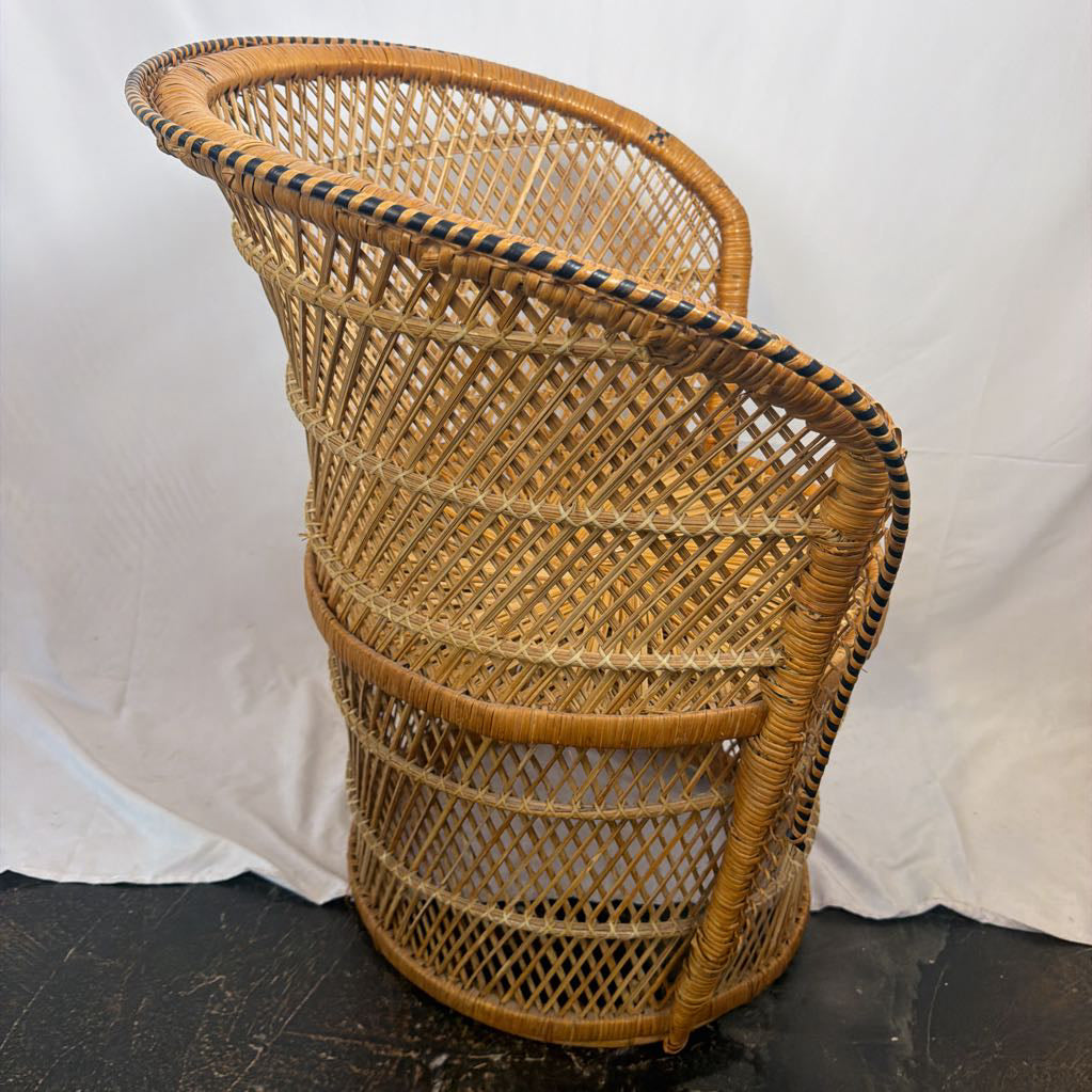 A natural rattan wicker chair with a curved back and seat, featuring a black and beige striped trim along the rim, set against a white fabric backdrop.