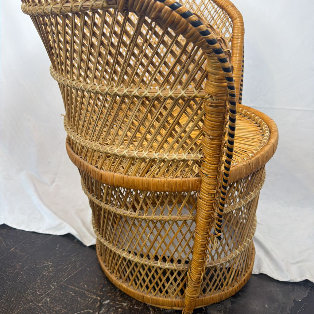 A natural woven rattan chair with a tall curved backrest, oval seat, and a black-and-tan striped hanging loop, photographed against a white draped backdrop.