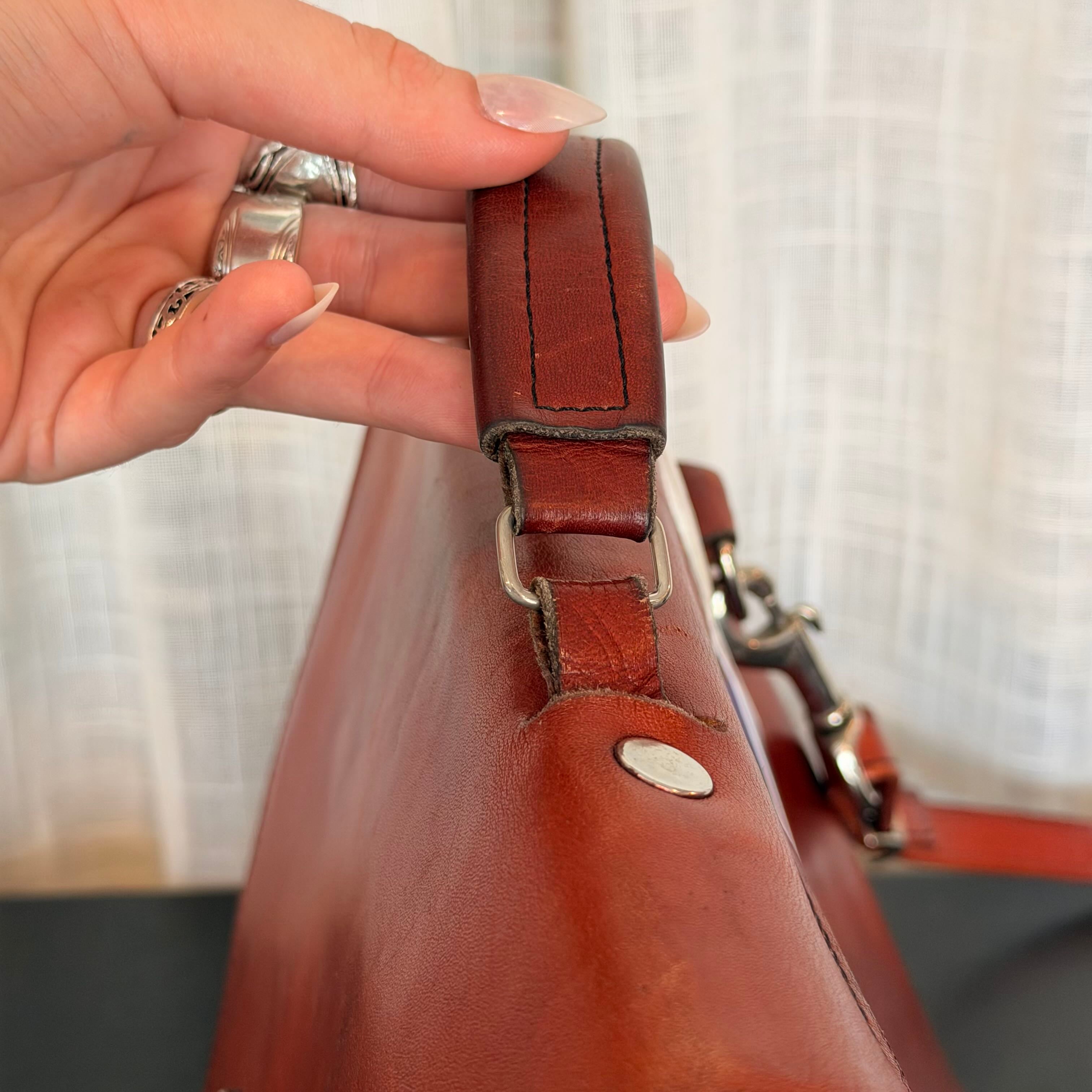 A close-up of a person's hand holding a brown leather bag handle, showing the texture of the leather and silver-tone hardware against a soft white curtain background.