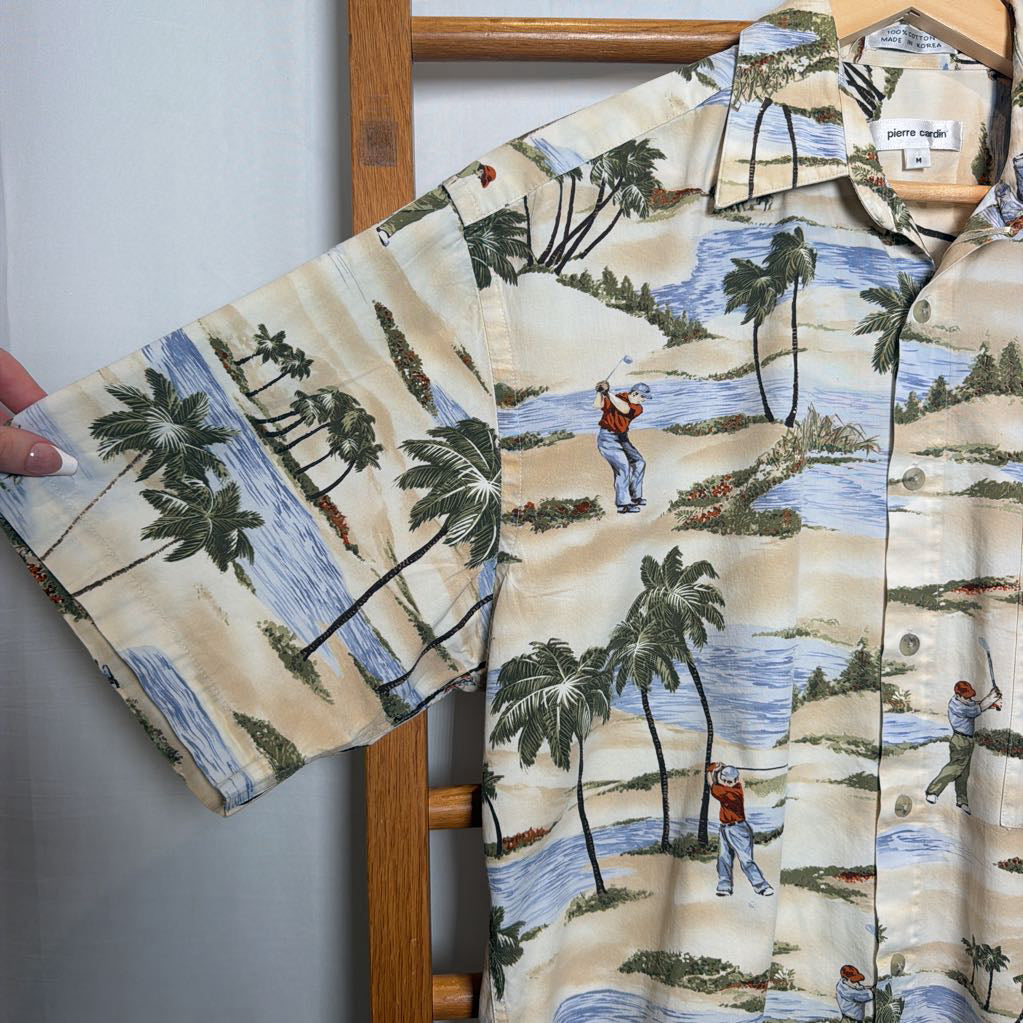 A short-sleeve button-up shirt with a tropical golf course print, featuring palm trees, blue water, and golfers on a sandy beige background. The shirt is displayed on a wooden hanger on a wooden chair, with a hand holding out the sleeve to show the fabric.