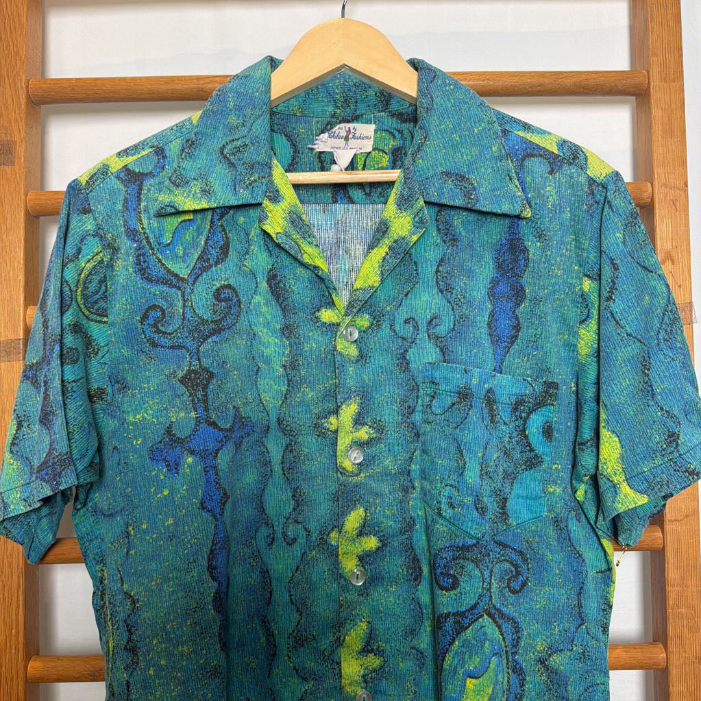 A short-sleeve button-up shirt with a vibrant blue, green, and yellow abstract pattern, hanging on a wooden hanger against a wooden slatted background.