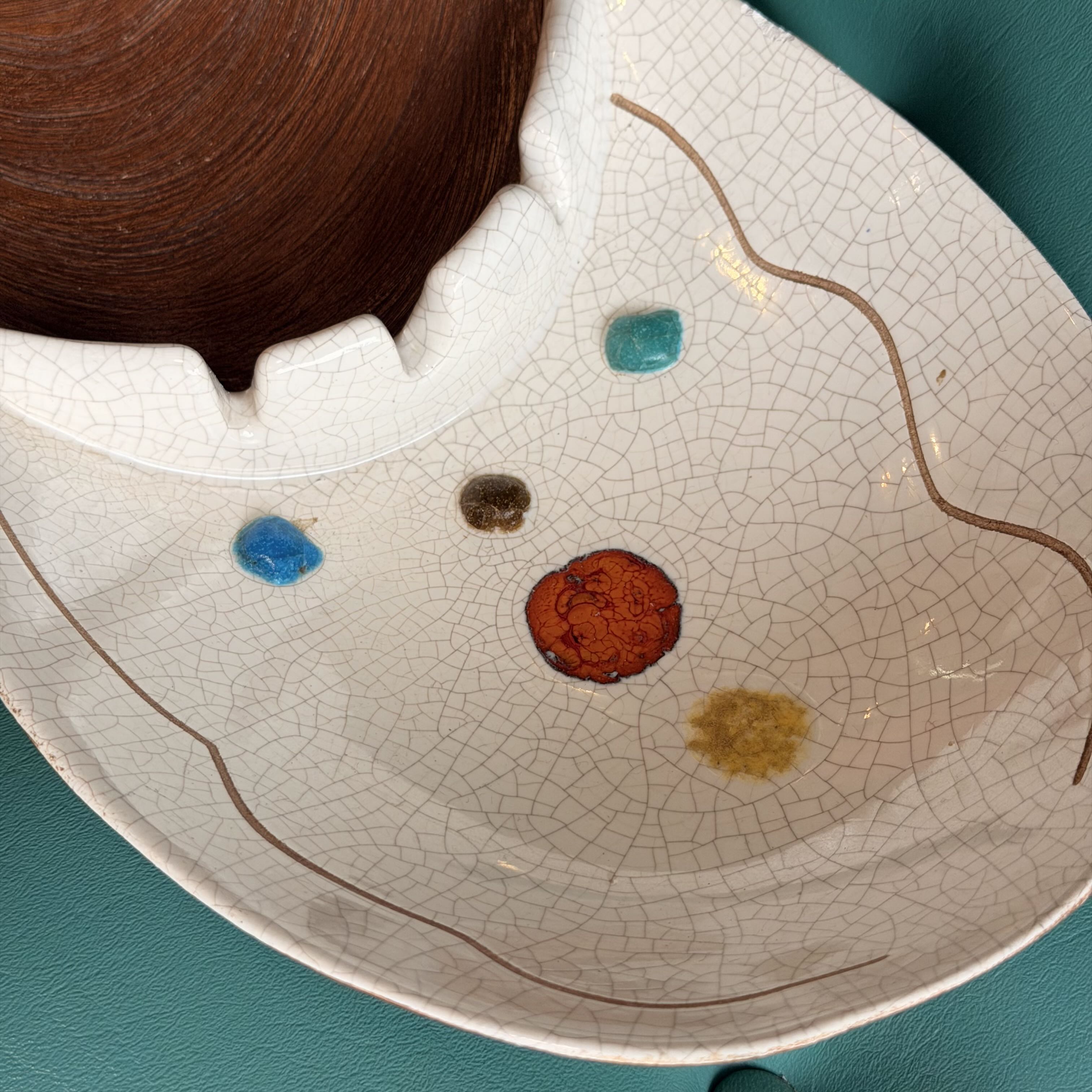 A close-up of a white crackle-glazed ceramic dish with a scalloped rim, decorated with small colorful spots and thin brown wavy lines, set against a teal background.
