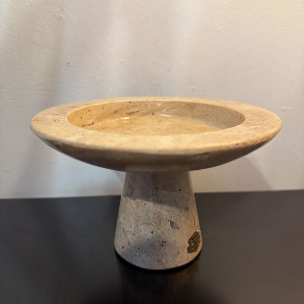 A small stone pedestal dish or candle holder made of natural beige and gray travertine, set on a dark reflective surface against a light gray wall.