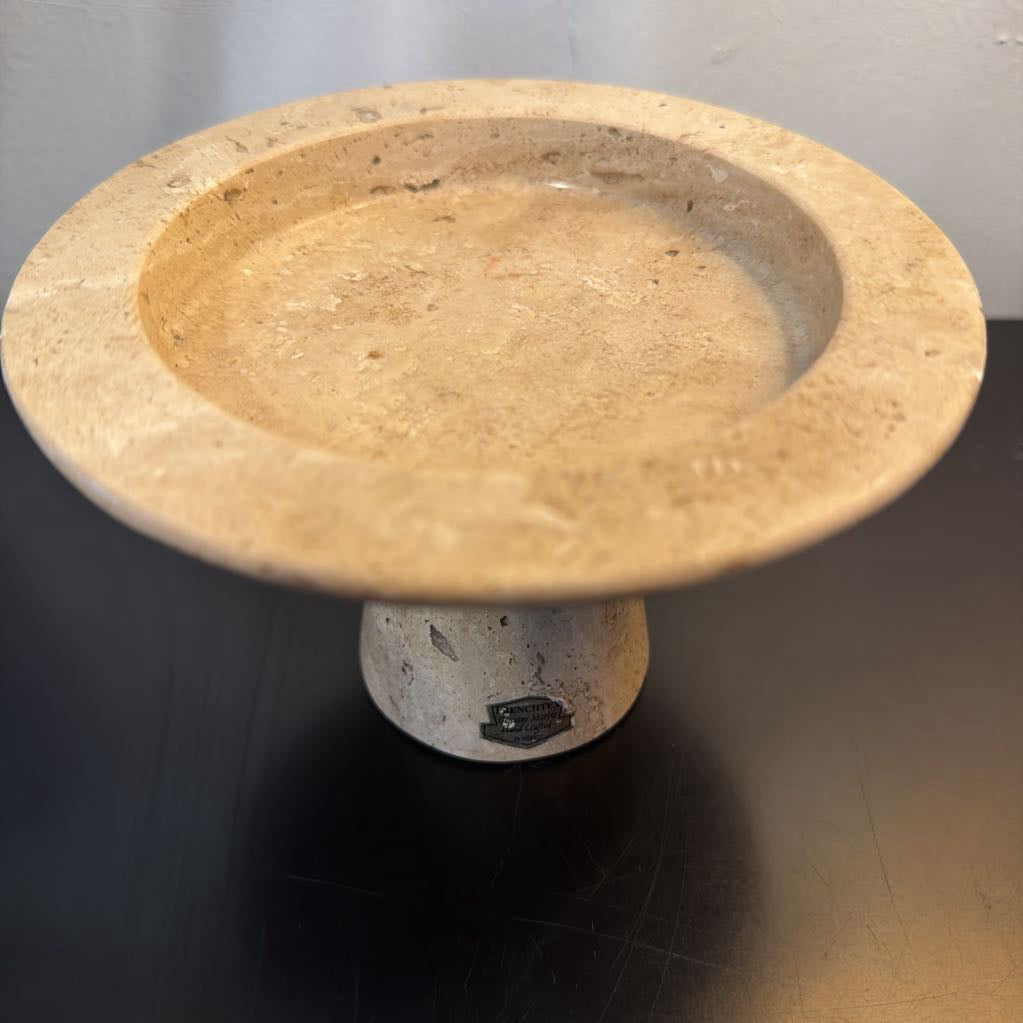 A round stone pedestal plate or dish with a wide shallow top and a short cylindrical base, shown on a dark reflective surface against a light gray wall.