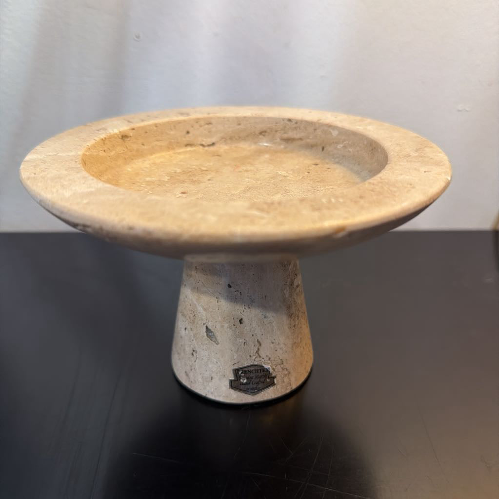 A round stone pedestal planter or decorative stand made of beige travertine or similar natural stone, shown on a dark reflective surface against a light gray wall.