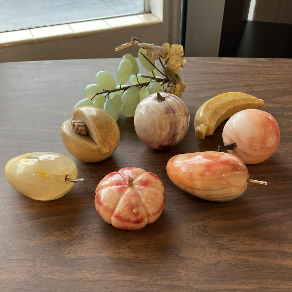 A set of decorative faux fruit ornaments arranged on a dark wooden table, including a yellow banana, green grapes, and marbled apples and pears in soft peach, cream, and pink tones.