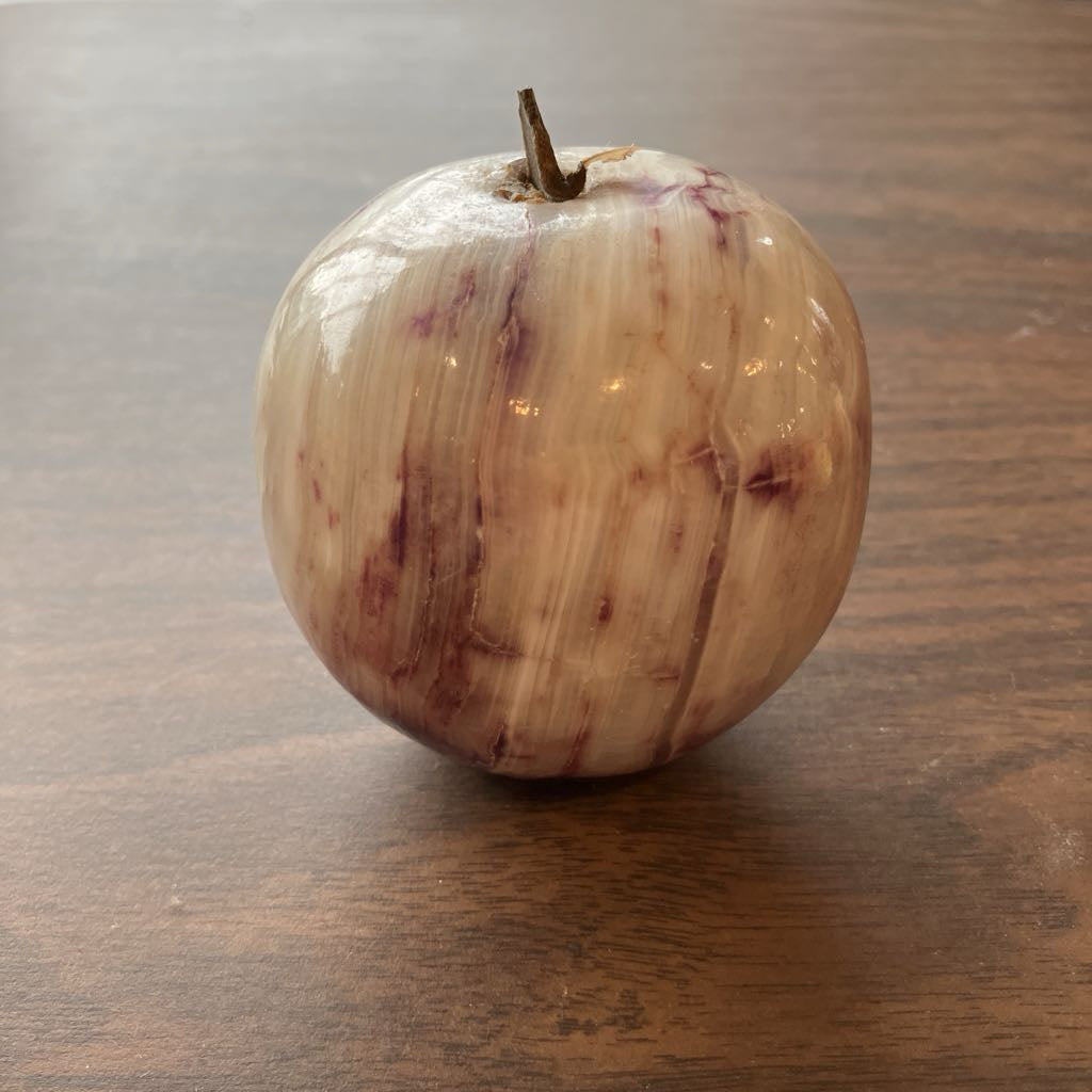 A carved decorative apple-shaped object with a natural wood-grain pattern in beige, cream, and reddish-brown tones, featuring a small stem at the top, set on a dark wooden tabletop.