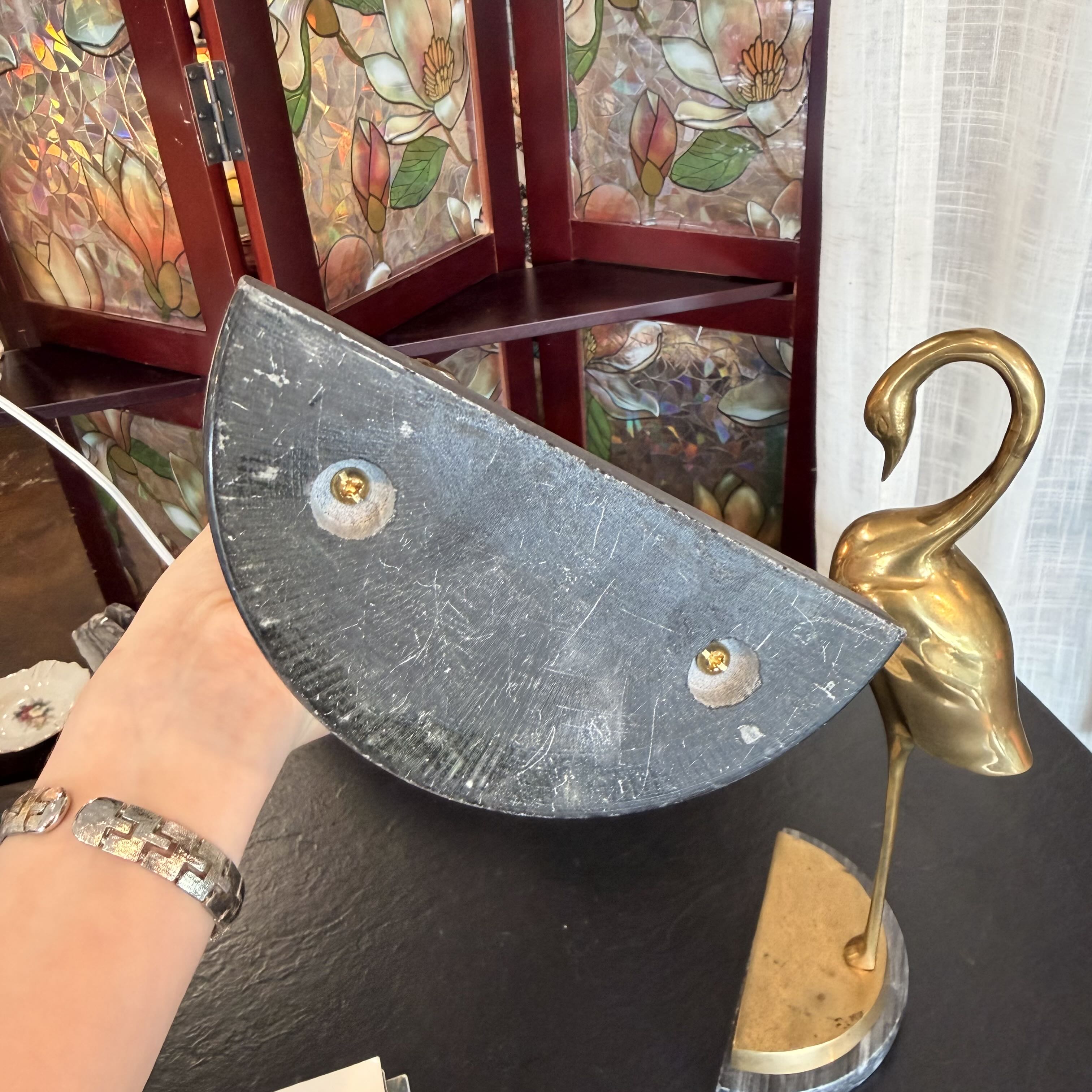 A hand holds a dark gray, semi-circular stone or metal wall plaque with two brass screws, next to a gold-toned flamingo figurine on a black table, with a stained-glass screen in the background.