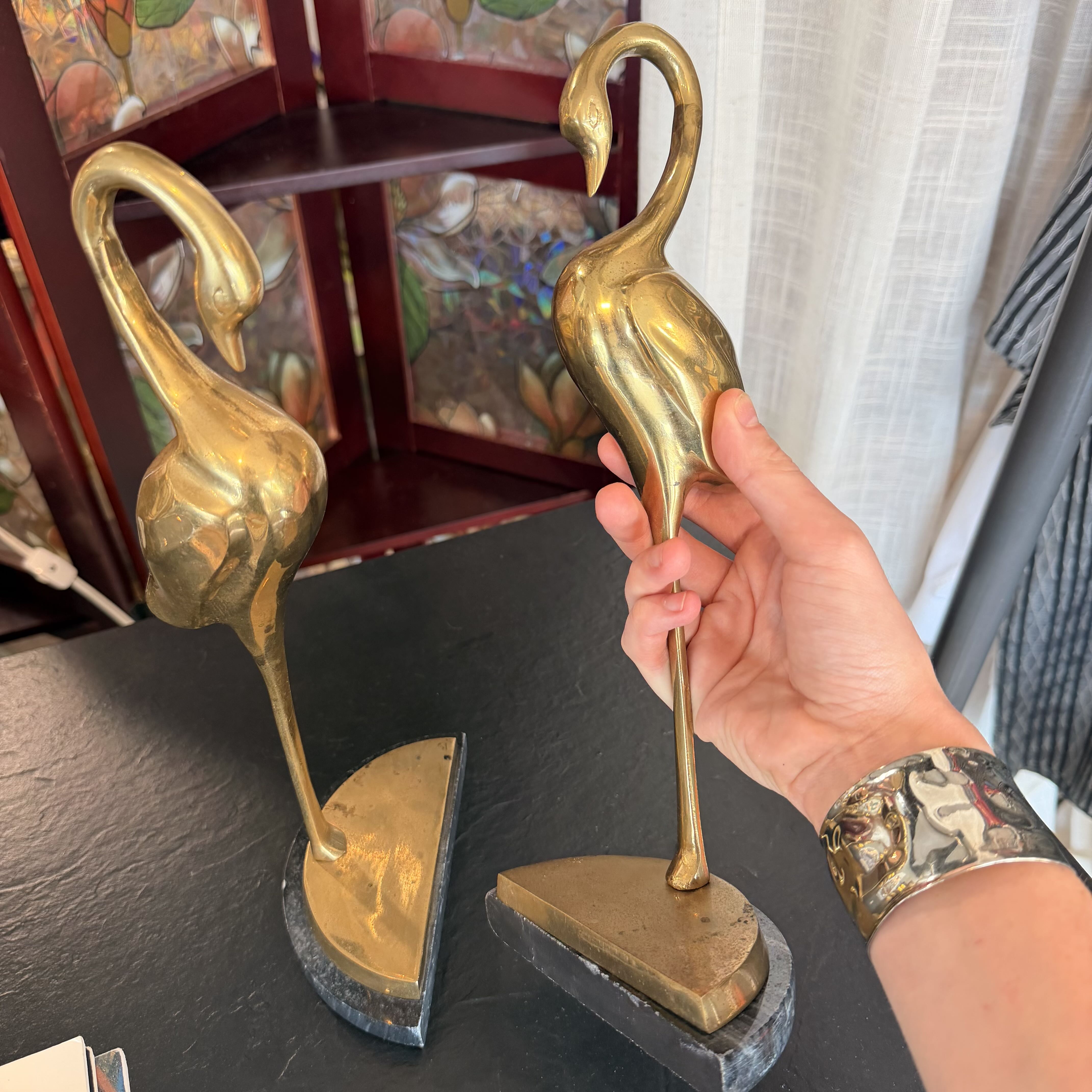 A pair of vintage-style gold-toned metal flamingo bookends on a black slate surface, with a hand holding one of the bookends to show its scale and finish.