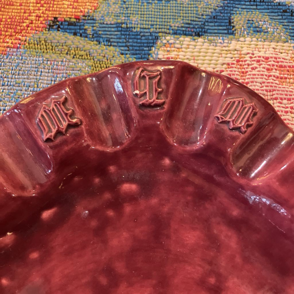 A close-up of a glossy, deep burgundy or maroon ceramic dish with embossed Gothic letter motifs along its scalloped rim, set against a colorful woven textile background.