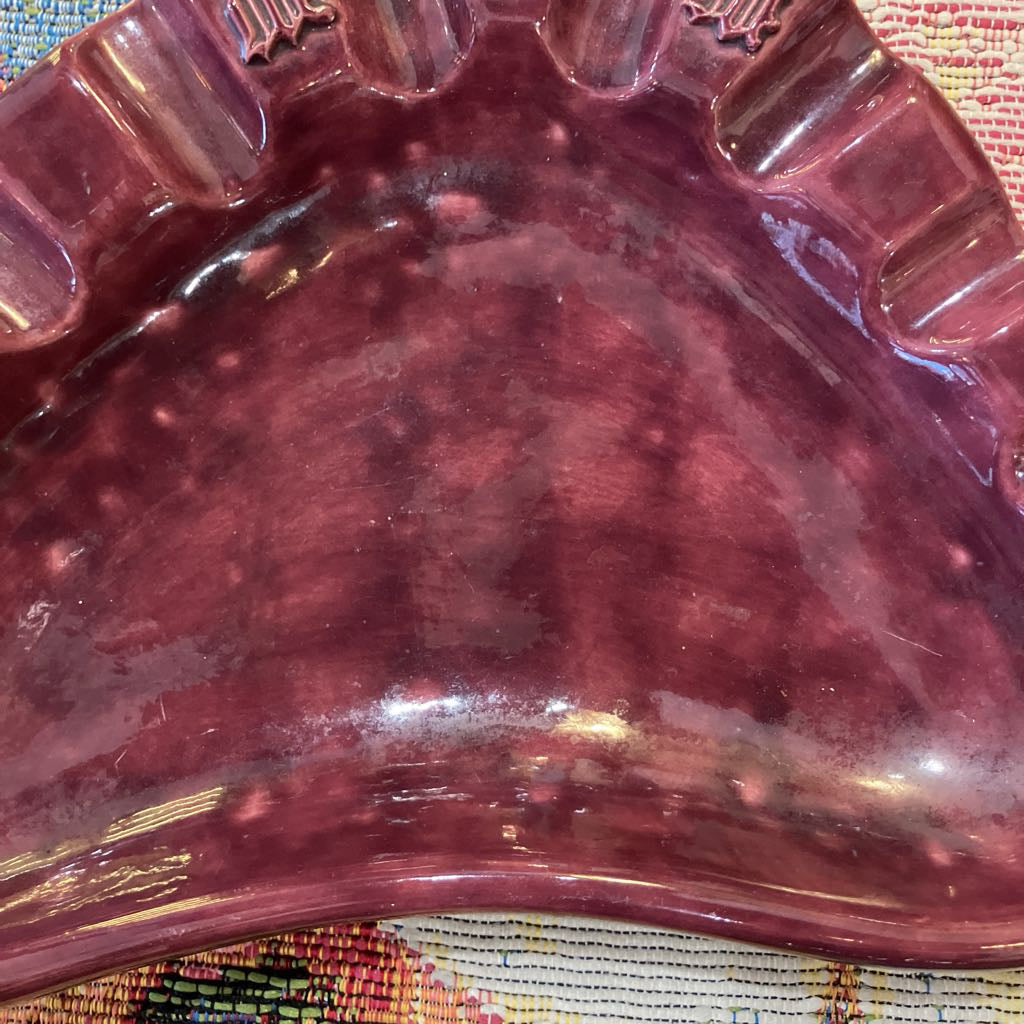 A close-up of a glossy, deep burgundy or maroon ceramic dish with a scalloped rim, resting on a textured multicolored woven fabric surface.