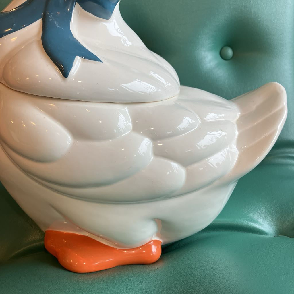 A glossy white ceramic duck-shaped container or figurine with a blue beak detail and orange feet, photographed close up against a teal tufted surface.