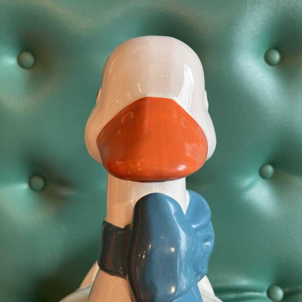 A glossy ceramic duck figurine shown from the back, featuring a white body, orange beak, and blue bow tie, set against a teal tufted leather background.