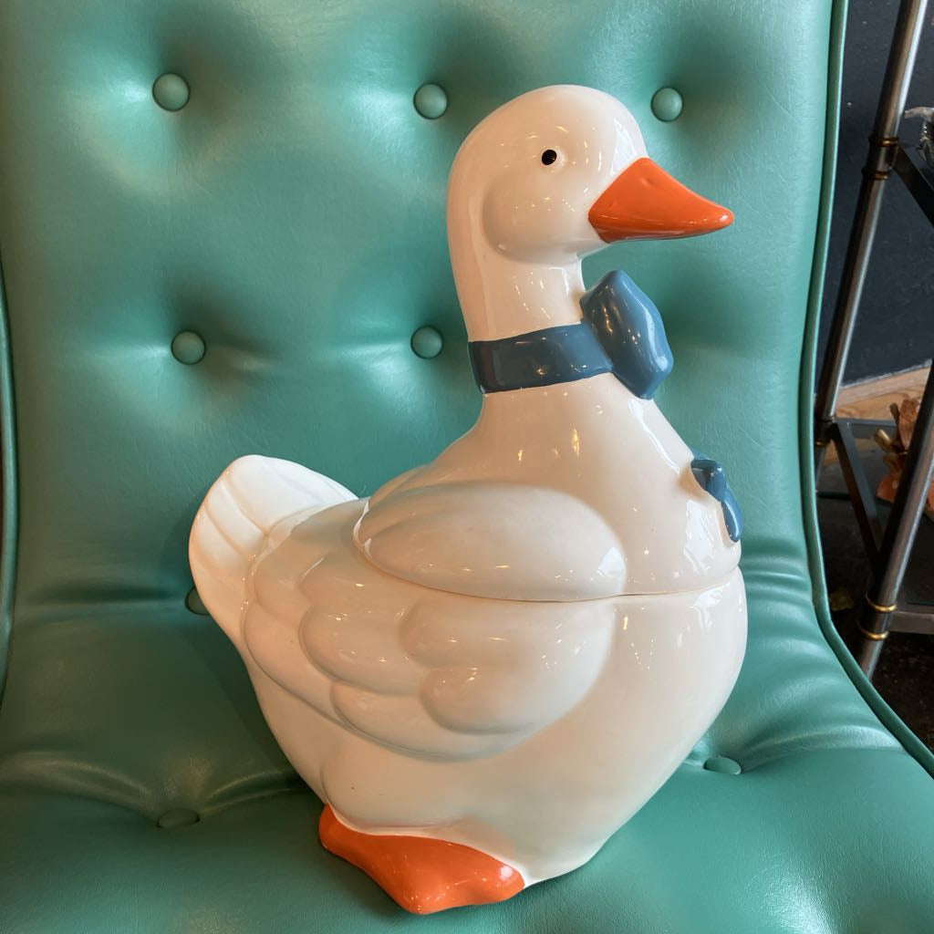 A glossy white ceramic duck figurine with an orange beak and feet, wearing a blue bow tie, displayed on a teal tufted chair.