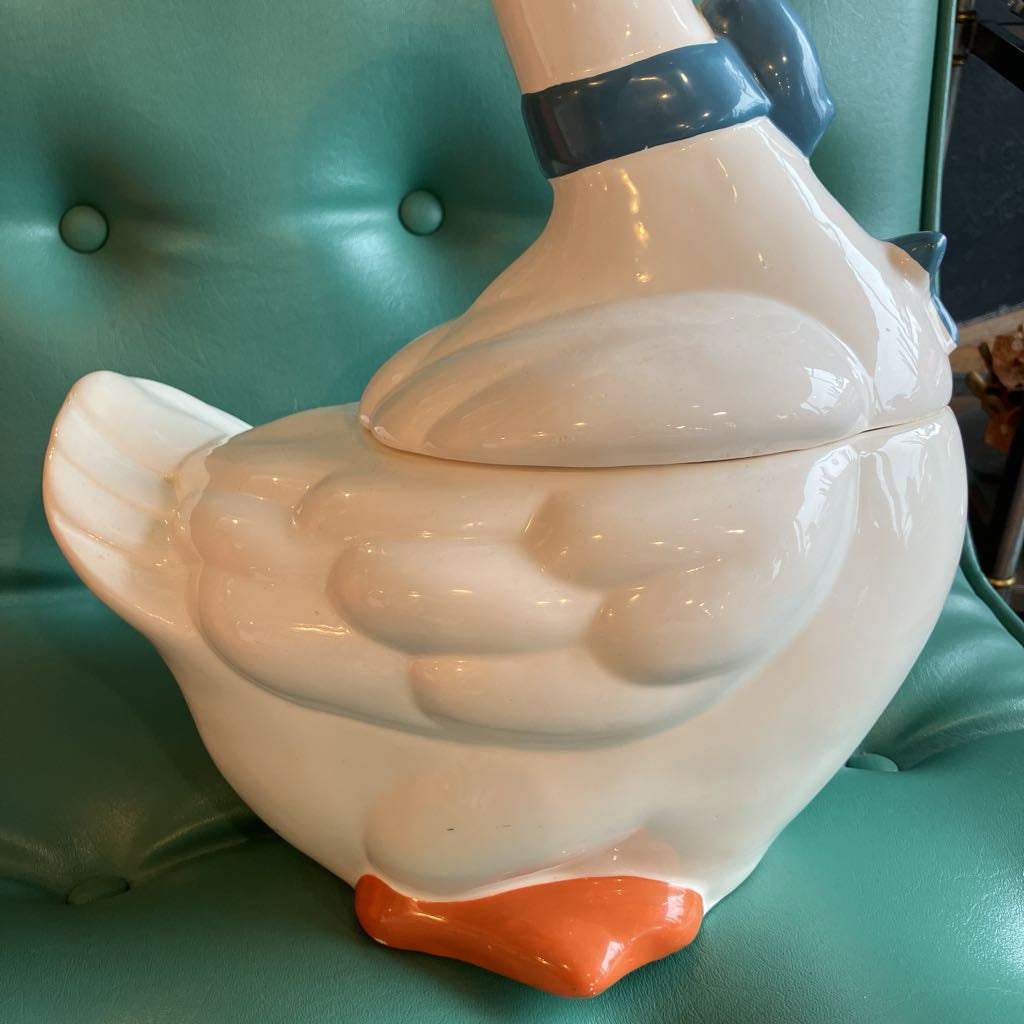 A glossy ceramic duck-shaped container or decorative piece with a white body, dark blue neck band, and orange beak, placed on a teal tufted chair.