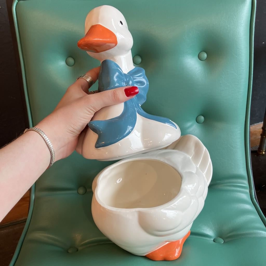A ceramic duck-shaped container or planter with a white body, orange beak and feet, and a blue bow detail, shown with a hand lifting the lid on a teal tufted chair.