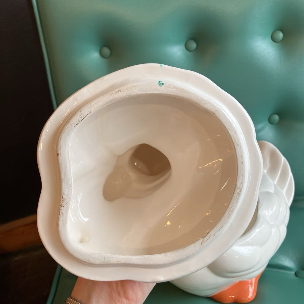 A white ceramic duck-shaped container or planter, shown from a top-down view inside the opening, held by a hand against a teal tufted leather background.