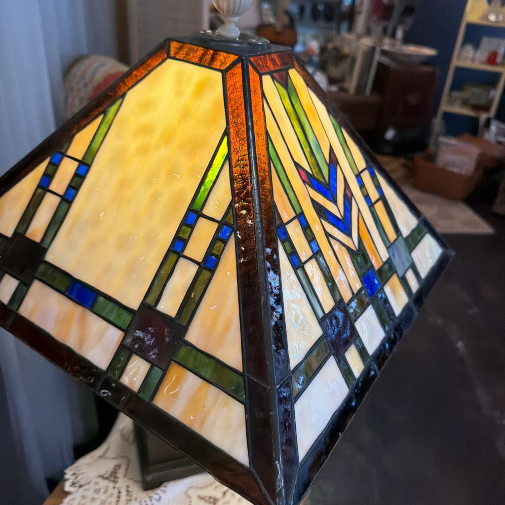 A stained glass table lamp shade with a geometric Art Deco design, featuring yellow, green, blue, and white glass panels held together by dark metal frames.