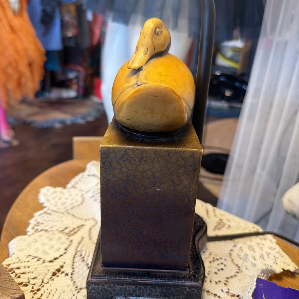 A small carved duck figurine in warm yellow and brown tones, displayed on a dark square pedestal with a crackled finish, set on a lace doily in a shop interior.