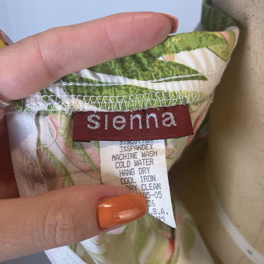 Close-up of a hand holding a patterned garment label that reads 'sienna' in white on a dark red tag. The fabric has a green, pink, and white tropical leaf print, and the hand has orange nail polish.