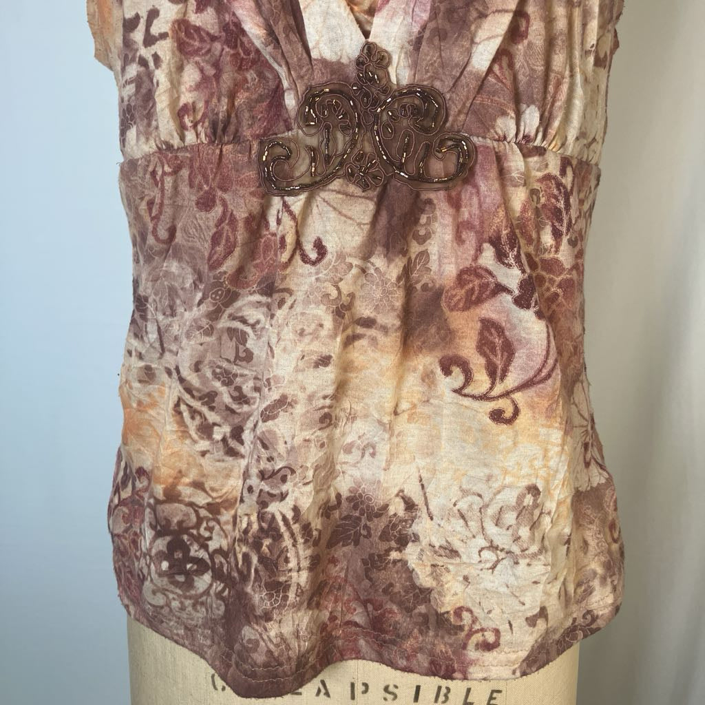 A close-up of a sleeveless blouse displayed on a dress form, featuring a brown and beige floral pattern with a decorative metallic applique at the bust.