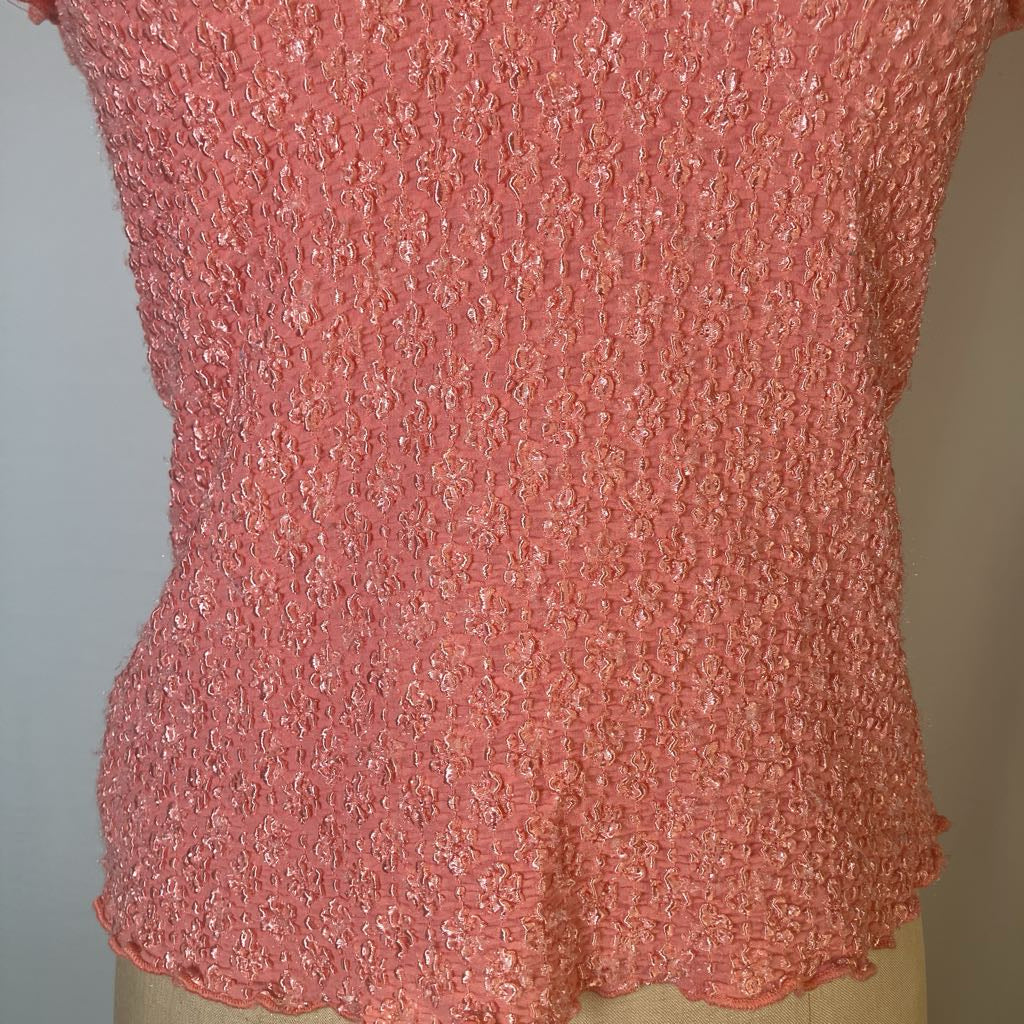 A close-up of a coral pink textured knit top displayed on a beige dress form, showing the detailed fabric pattern and scalloped hem.