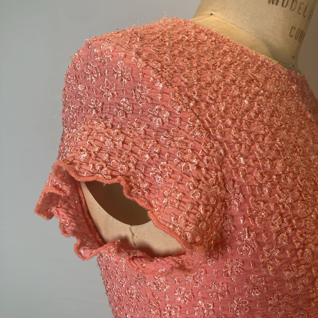A close-up of a textured coral pink dress or fabric on a mannequin torso, showing a ruffled sleeve and a cutout detail against a plain light gray background.