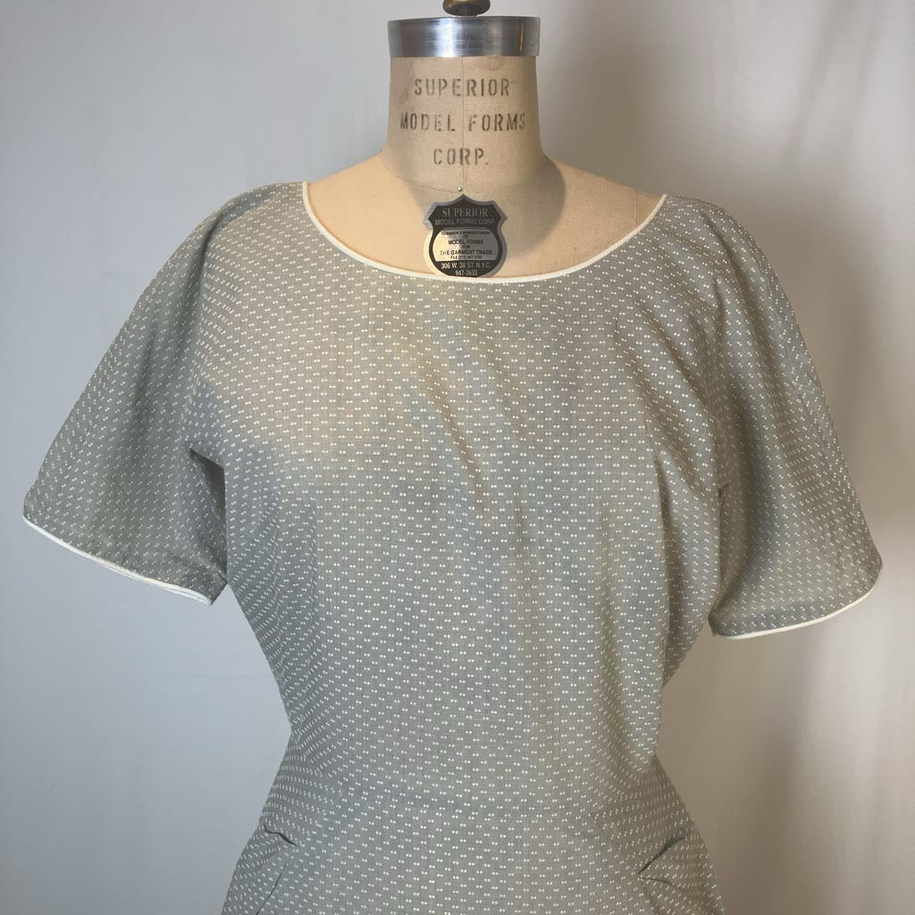 A short-sleeve blouse with a small white geometric pattern on a light gray base, displayed on a vintage mannequin torso. The blouse has a scoop neckline with white piping and short sleeves with white piping at the cuffs.