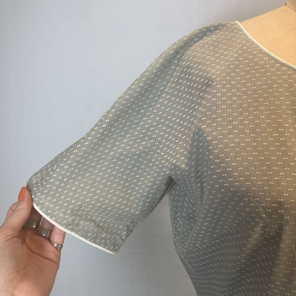 A close-up of a short-sleeve blouse on a mannequin or dress form, with a hand holding the sleeve to show the fabric and cuff. The blouse has a sheer, semi-transparent texture with a small white geometric dot pattern over a muted gray-green base, finished with a white piped cuff.