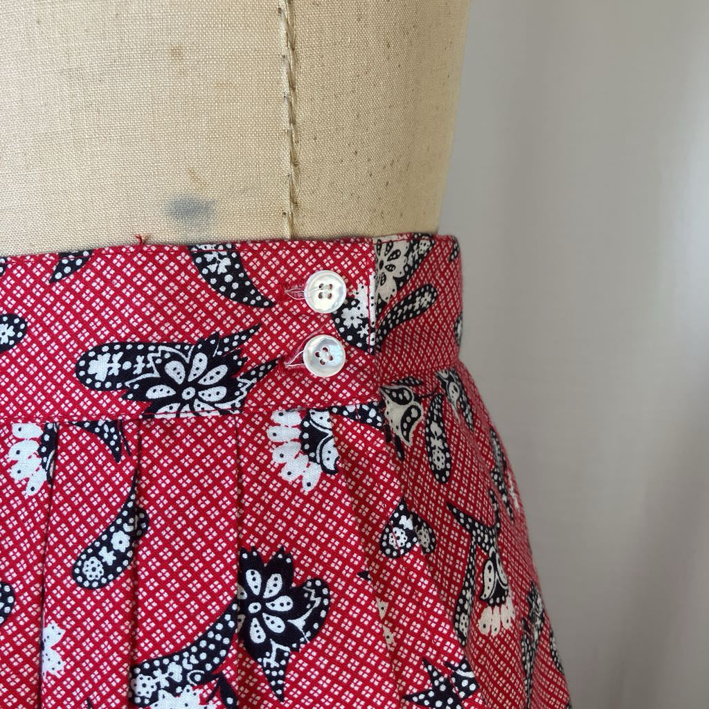 Close-up of a red patterned skirt or fabric panel on a beige dress form, featuring black and white paisley-style motifs and two white buttons near the waistband.