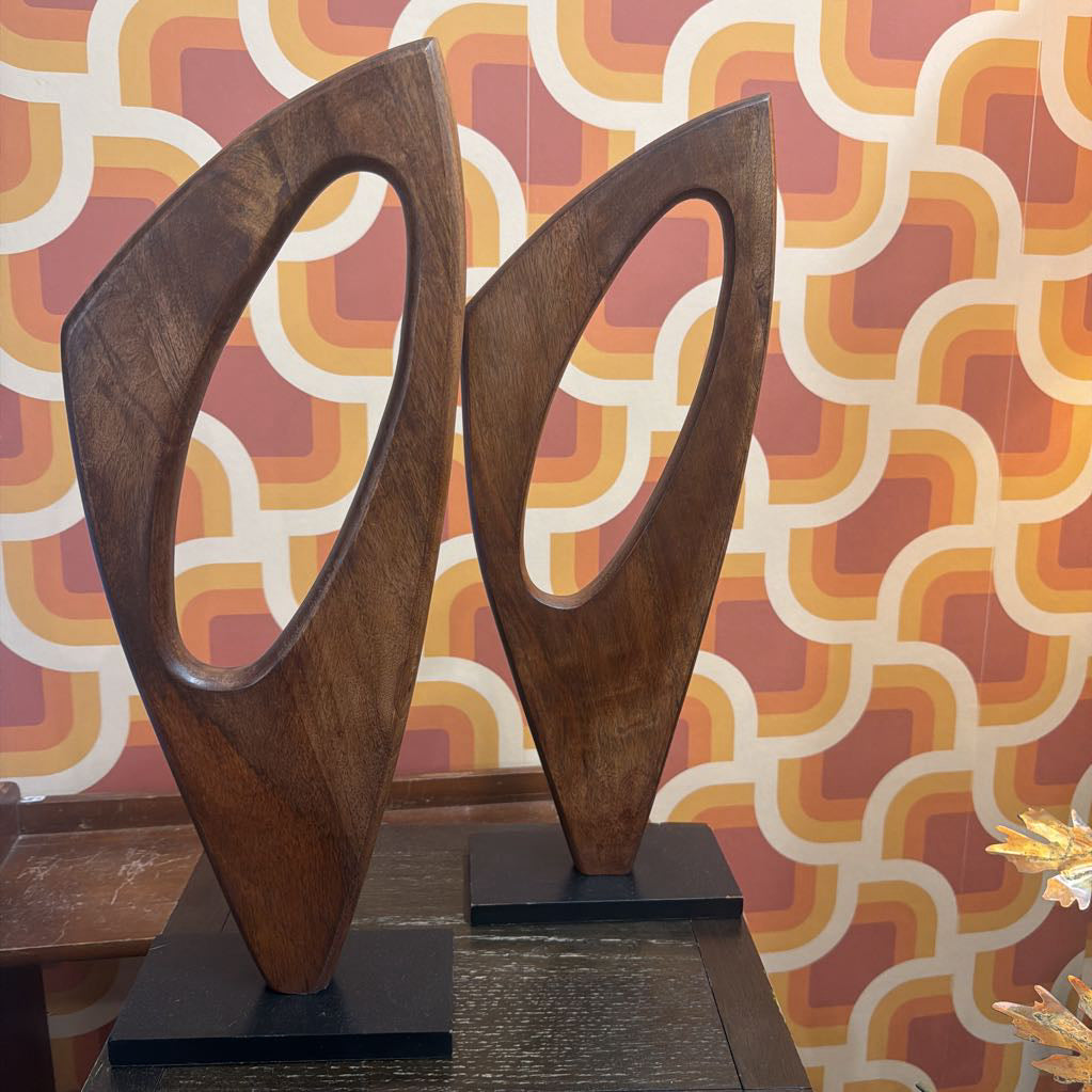 Two modern wooden abstract sculptures with curved, looped shapes mounted on black square bases, set against a retro patterned wallpaper background.