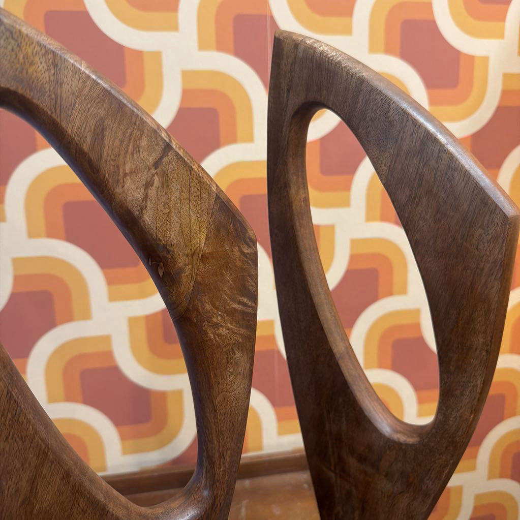 Close-up of two dark brown wooden chair backs with open circular cutouts, set against a retro patterned wallpaper in orange, yellow, and cream.