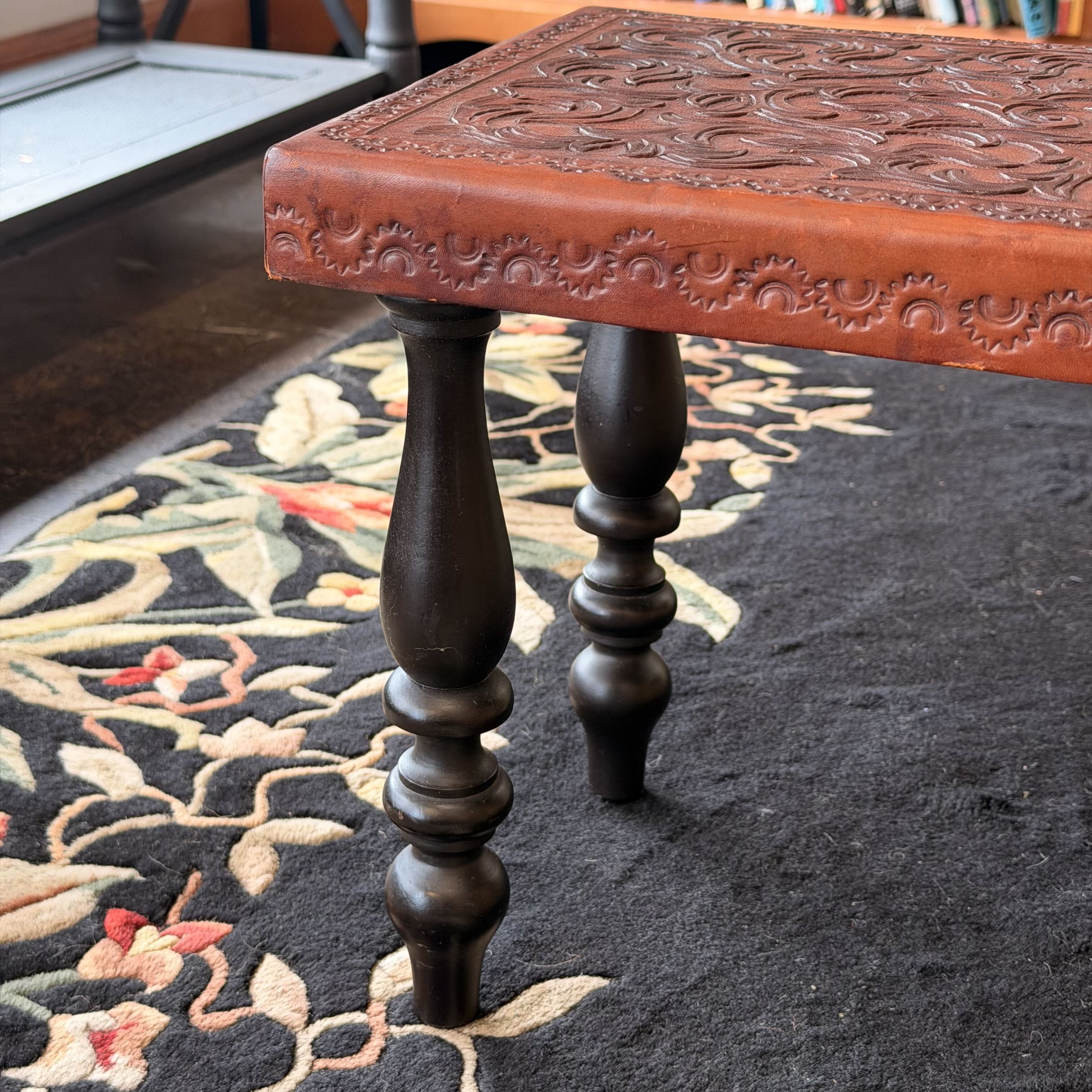 A close-up of a carved wooden stool or bench with a reddish-brown ornate top and dark turned legs, placed on a black floral rug in an indoor setting.