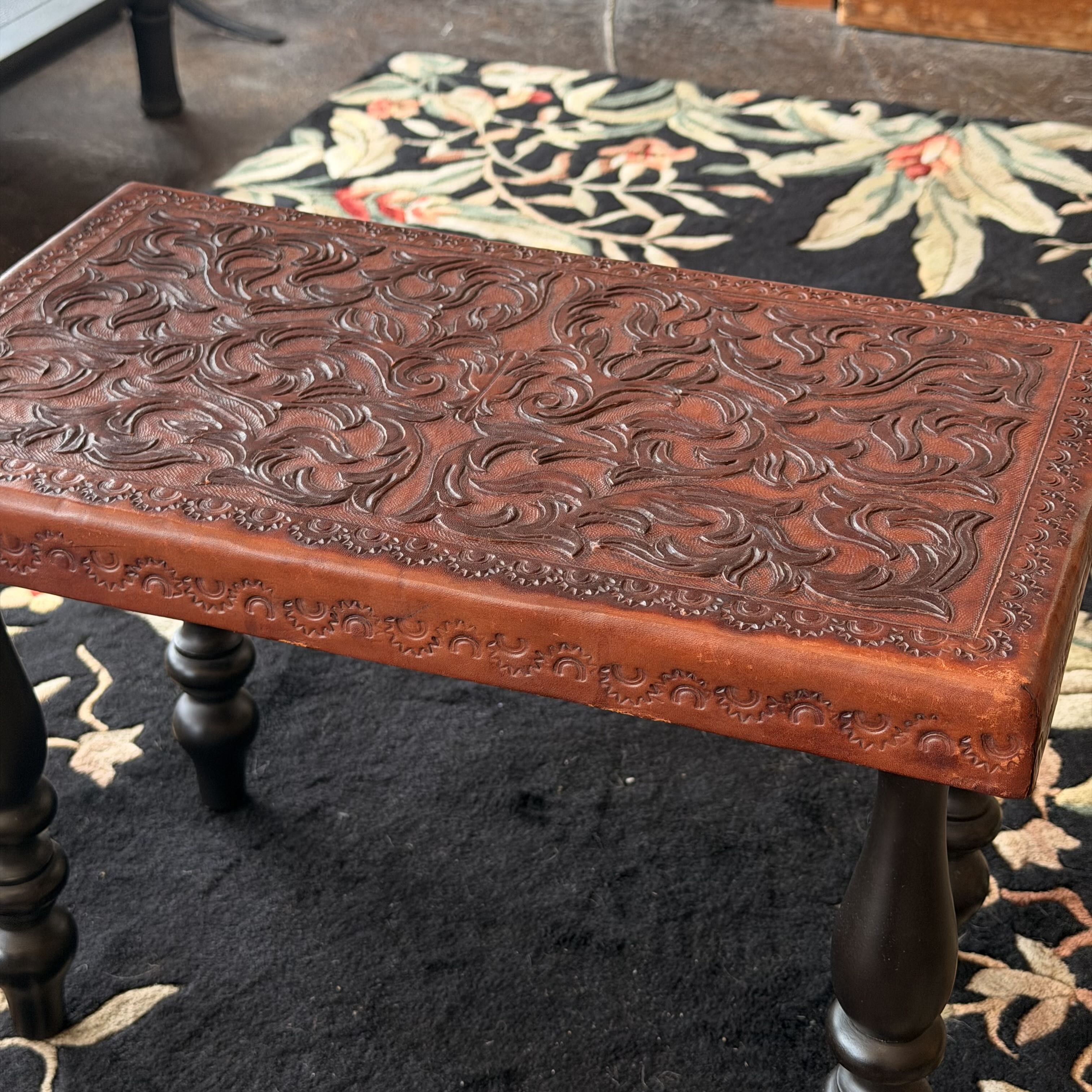 An ornate carved wooden bench with a reddish-brown carved top and dark legs, placed on a black floral rug.