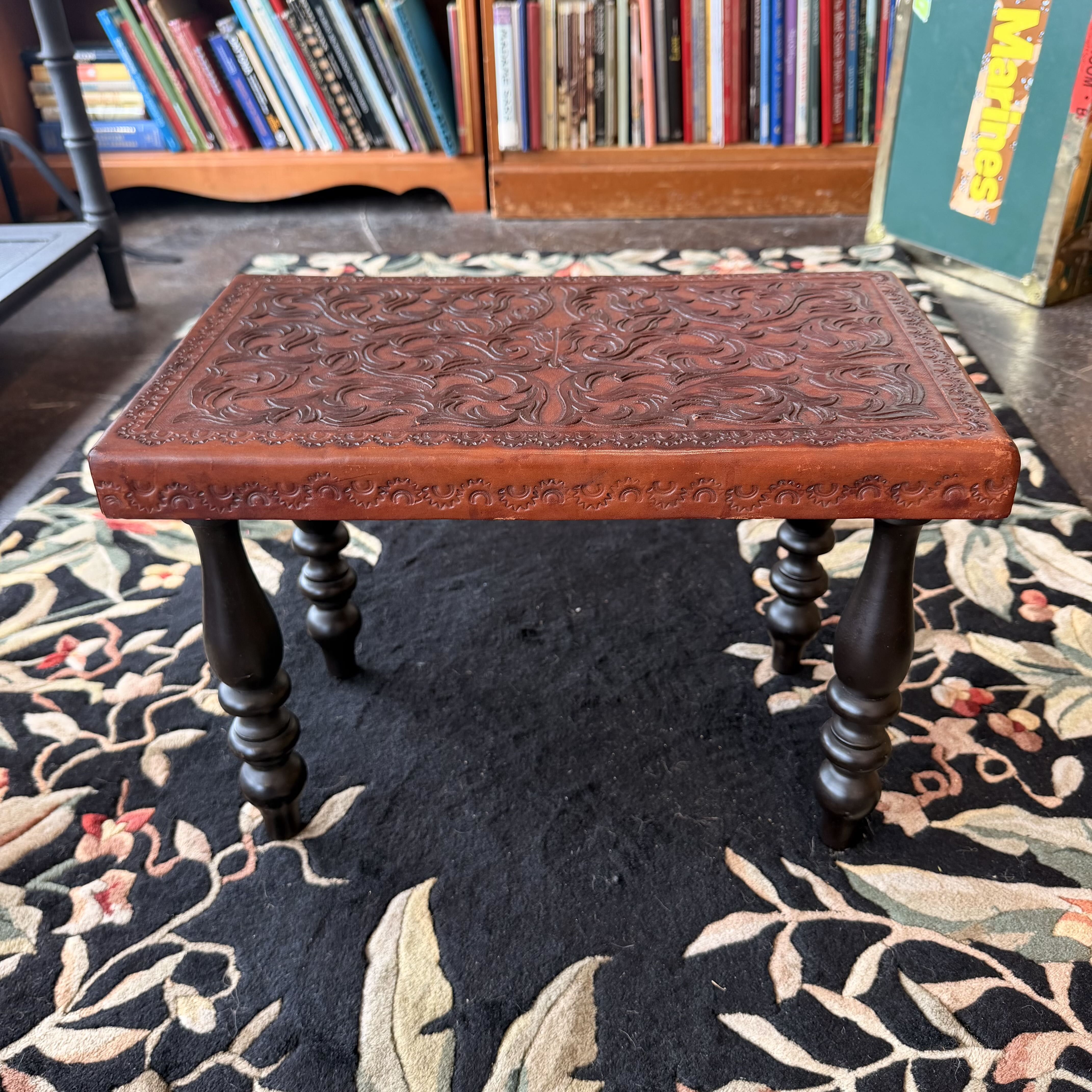 A small carved wooden bench or stool with an ornate embossed floral pattern on the seat and dark turned legs, placed on a patterned rug in front of a bookshelf.