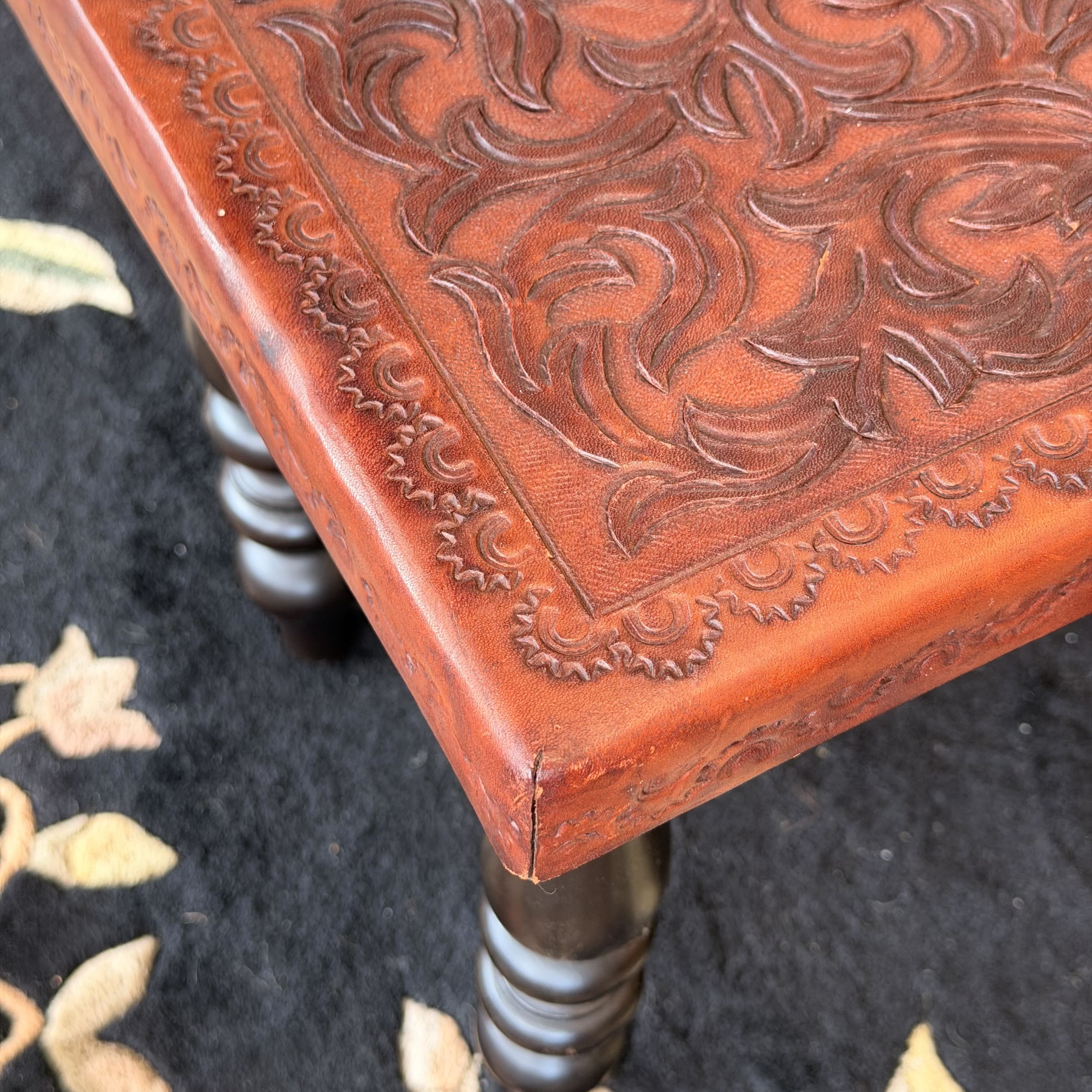 Close-up of a carved reddish-brown wooden stool corner with ornate embossed scrollwork and dark turned legs on a dark textured rug.