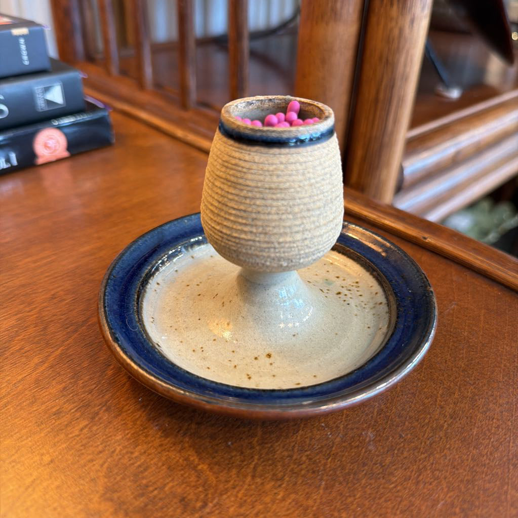 A handmade ceramic candle holder with a textured beige body and a deep blue glazed rim, shown on a wooden table with a blurred background.