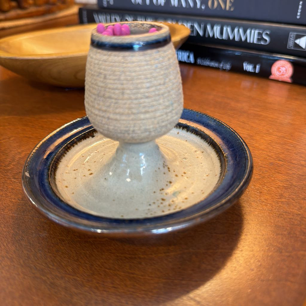 A handmade ceramic candle holder with a textured beige body, a dark blue glazed rim, and small pink wax drips on top, shown on a wooden table with books in the background.