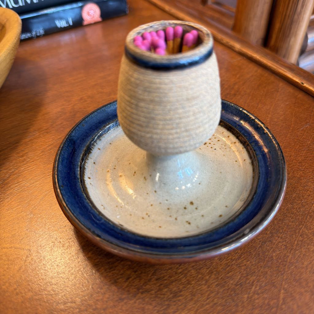 A handmade ceramic candle holder with a textured beige body, a dark blue glazed rim, and a matching round base, shown on a wooden table with a small set of pink matches inside.