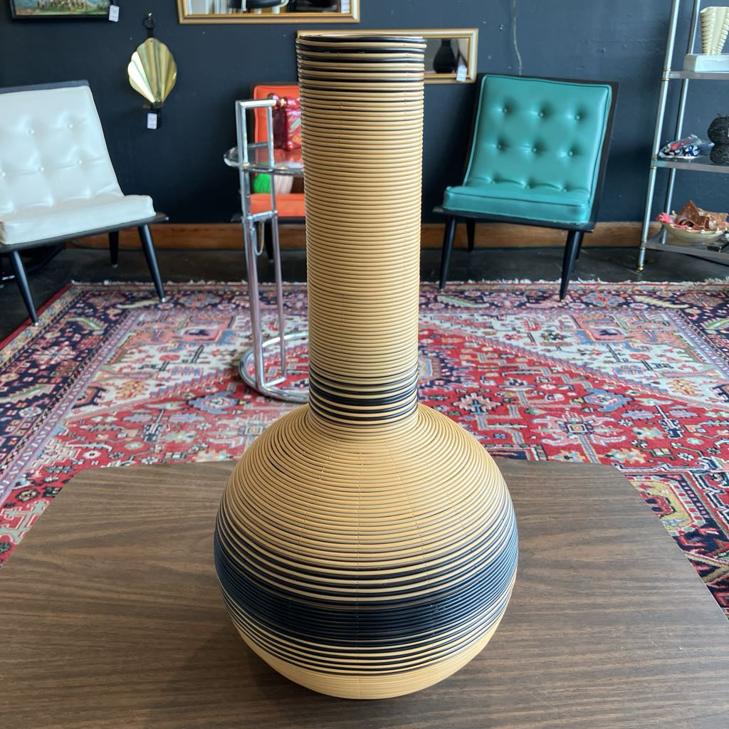 A tall, sculptural reed vase with a flared round base, made from stacked wood strips in a light tan color with dark charcoal horizontal bands, displayed on a wooden table in a room with patterned rugs and chairs.