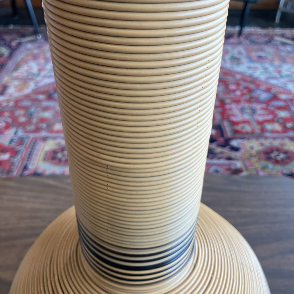 A close-up of a woven natural fiber lamp base, likely rattan or seagrass, with a flared circular bottom and a ribbed cylindrical upper section, set against a blurred indoor background with a patterned rug.