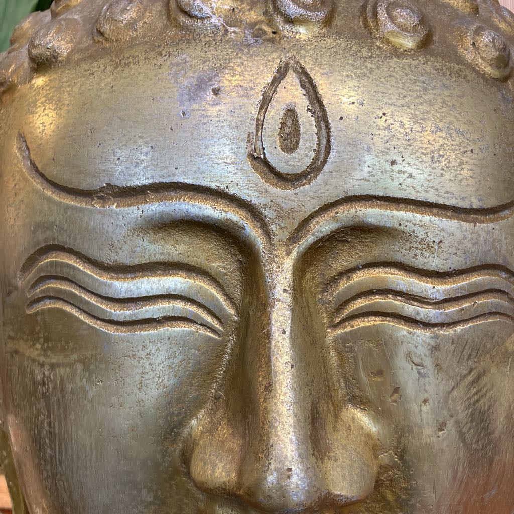 Close-up of a metallic Buddha face sculpture with a textured bronze finish, showing carved facial features and a teardrop-shaped forehead symbol.
