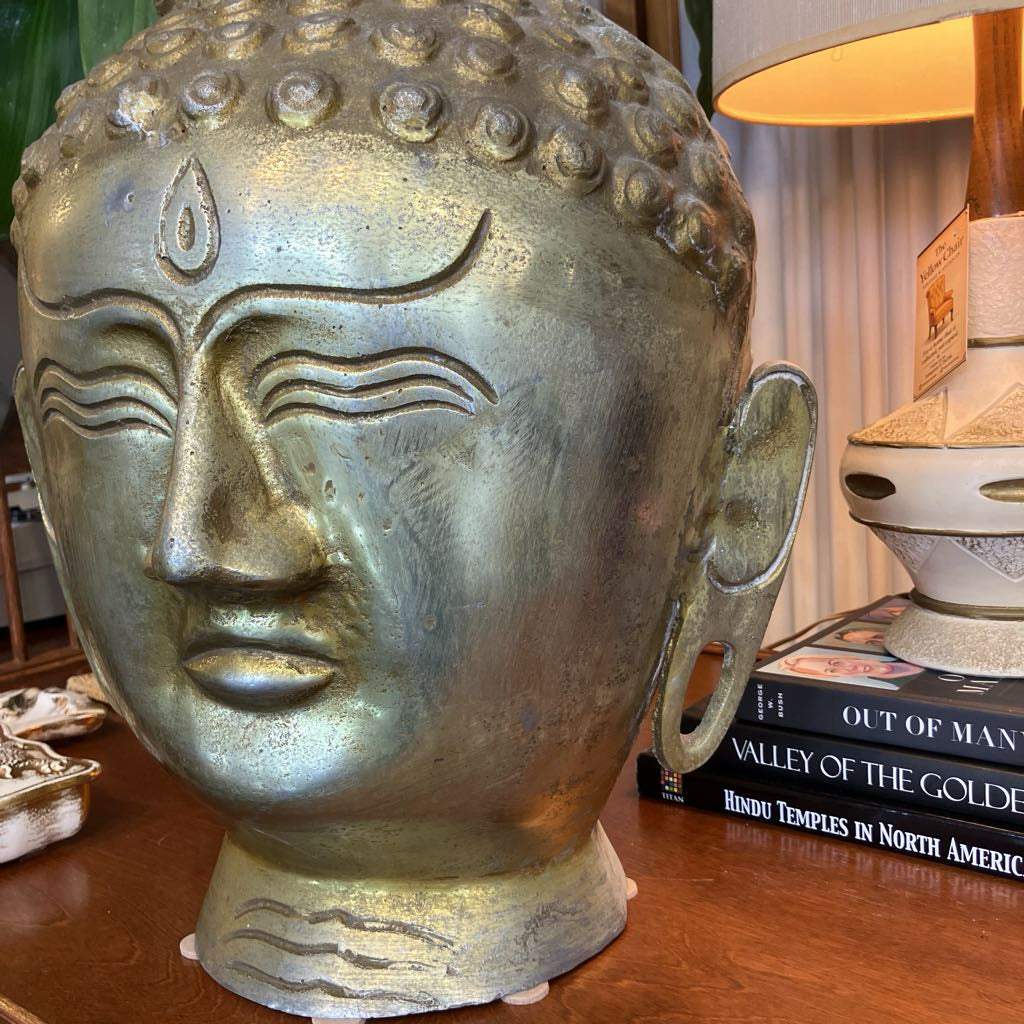 A large metallic Buddha head sculpture with a weathered bronze finish, displayed on a wooden table next to a stack of books and a lamp.