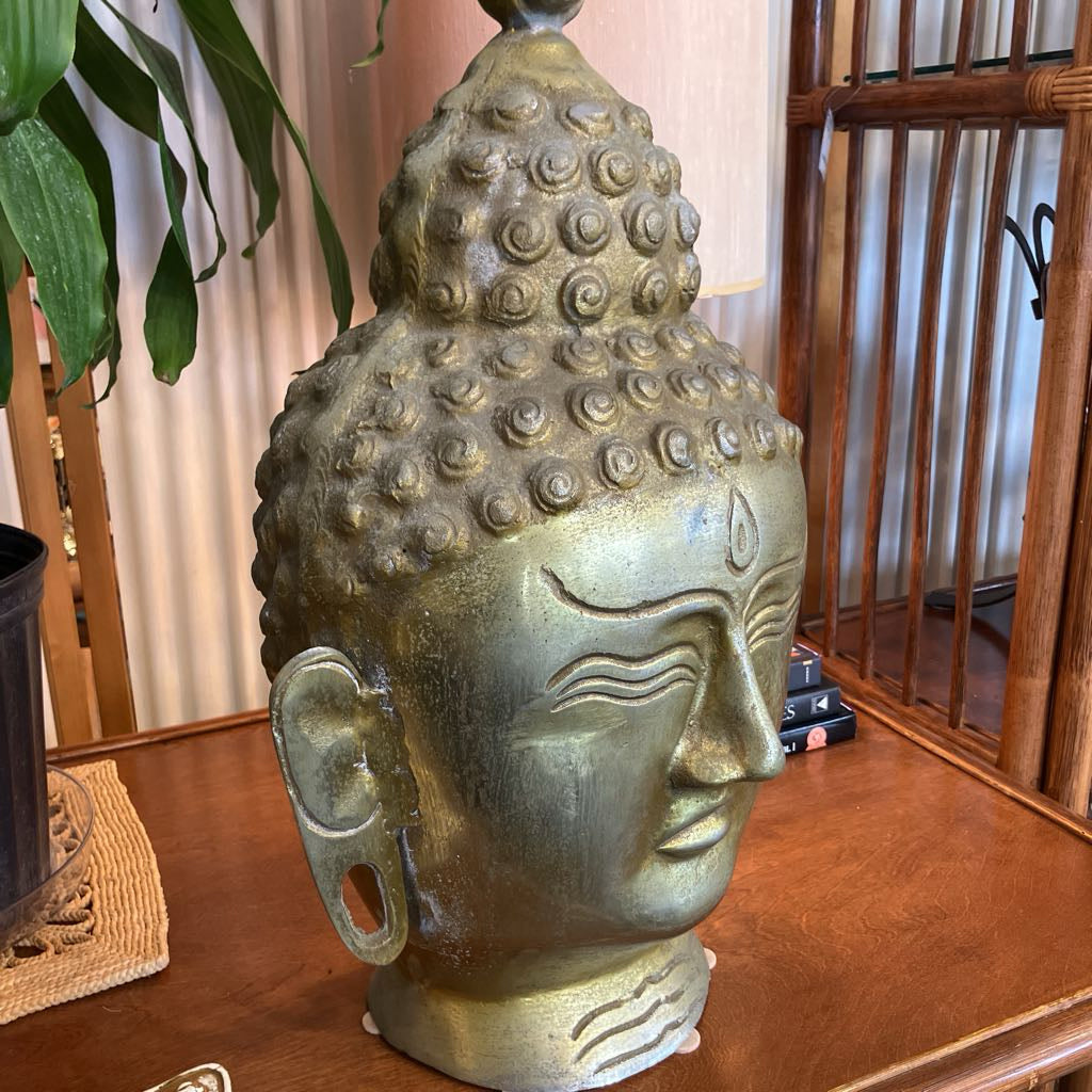 A large decorative Buddha head sculpture with a textured metallic finish, displayed on a wooden table in an indoor setting with a plant and wooden furniture in the background.