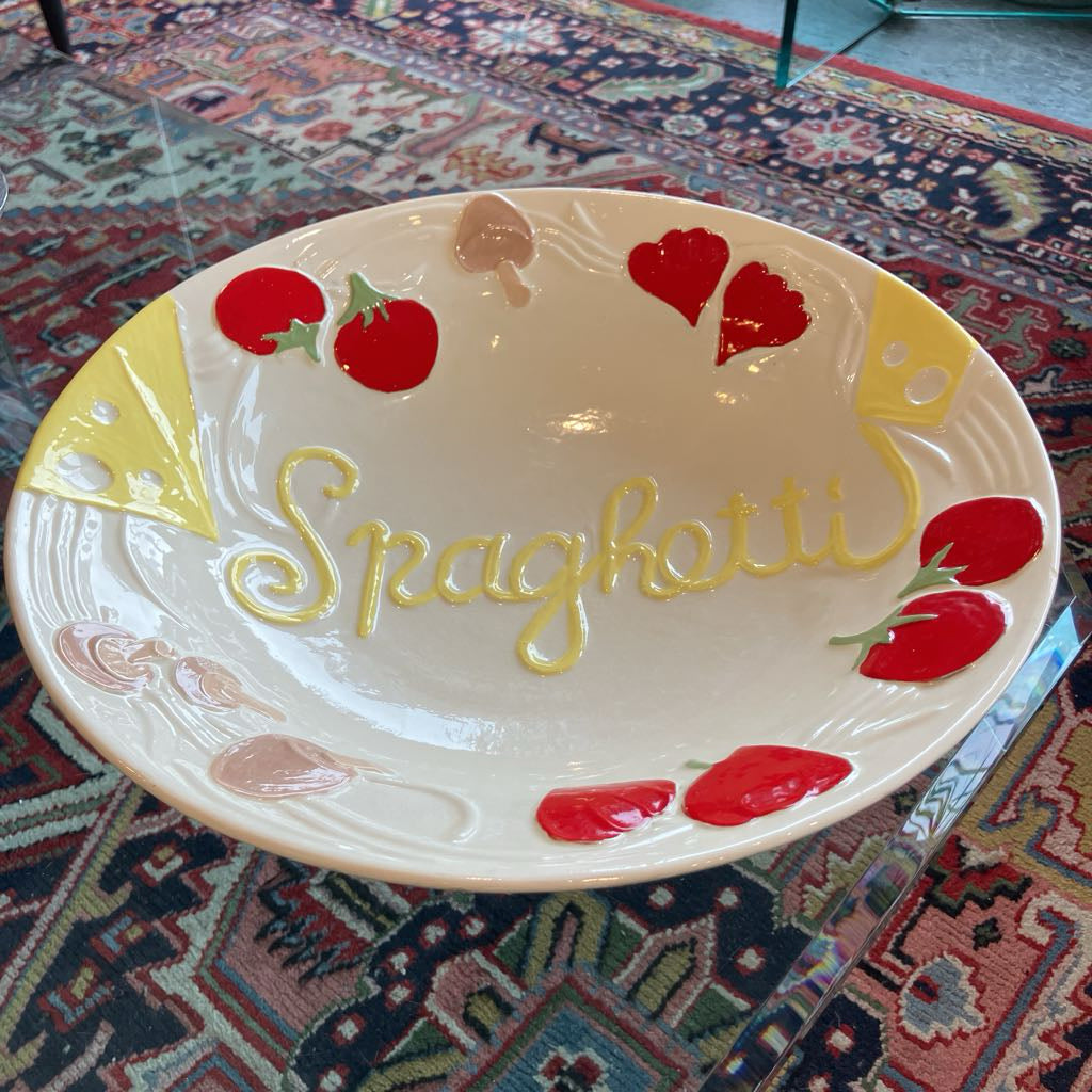 A ceramic serving platter shaped like a spaghetti dish, featuring the word 'Spaghetti' in yellow script, red tomato motifs, and yellow cheese wedge designs, displayed on a patterned rug.