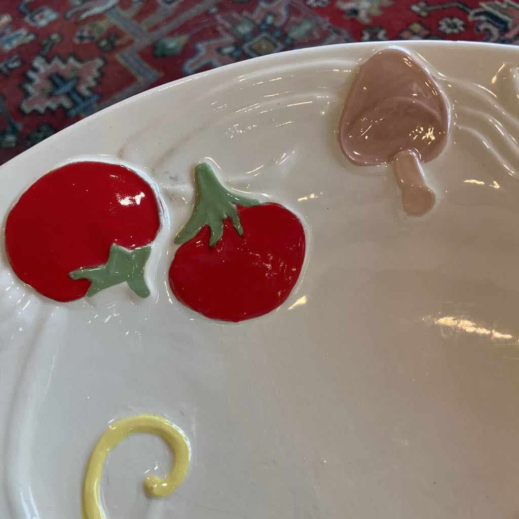 A close-up of a white ceramic plate featuring raised, sculpted decorative elements including two red tomatoes with green tops and a beige mushroom, with a small yellow curl at the bottom.