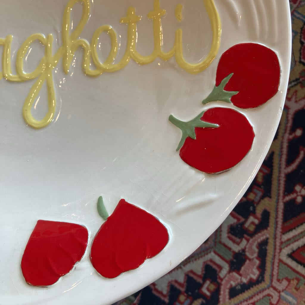 A close-up of a white ceramic plate with decorative yellow script lettering and red tomato motifs with green stems, set against a patterned rug.