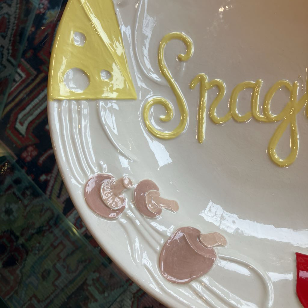 Close-up of a ceramic plate with raised pasta-inspired decorations, including yellow script lettering, a yellow cheese wedge motif, and pink pasta shapes on a glossy off-white surface.