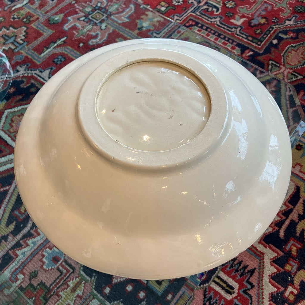 An off-white ceramic bowl shown from a top-down perspective, resting on a red and navy patterned oriental rug.